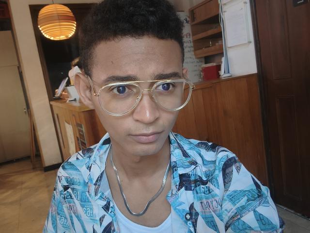 Um homem com cabelo preto encaracolado, usando óculos de aro dourado, camisa com estampa azul e colar de prata, olhando diretamente para a câmera com uma expressão neutra..