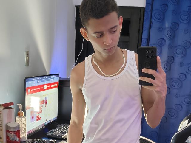 Raamking with short curly hair, wearing a white tank top, taking a mirror selfie, holding a black phone, standing in front of a computer and blue curtain.