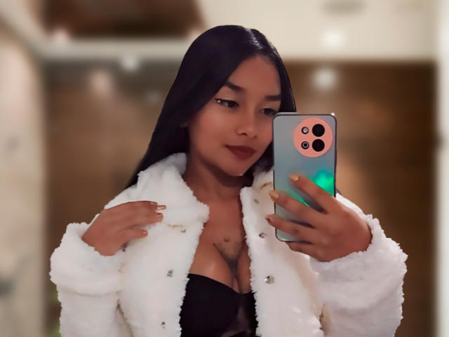 CloeMorgan with long black hair, wearing a fluffy white jacket, black lingerie, and holding a phone, showing her tits in a mirror selfie, smiling slightly.