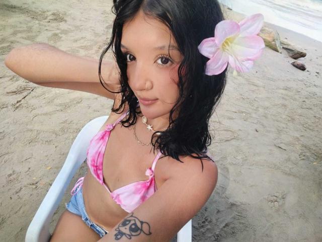PaulineRey with wet black hair, pink tie-dye bikini top, blue shorts, pink flower in hair, tattoo on right arm, sitting on white chair, sandy beach, looking up.