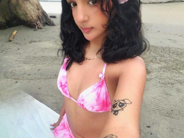 PaulineRey with wavy black hair, pink tie-dye bikini, small tits, tattoo on right arm, pink flower in hair, sitting on beach, tan skin, subtle smile, necklace.