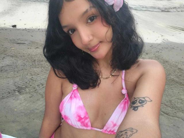 PaulineRey met golvend zwart haar, een roze en witte bikini, kleine borsten, een tatoeage op de rechterarm, een neusring, lachend, een roze haarspeldje, zittend op een zandstrand..