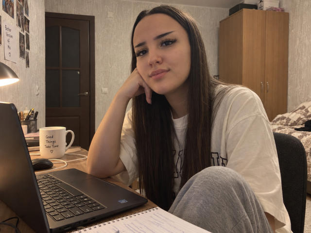 AdelleHoos, con lunghi capelli castani, una maglietta bianca e pantaloni grigi, seduta a una scrivania, con la testa appoggiata alla mano, un portatile davanti a lei, un leggero sorriso, un letto disordinato sullo sfondo..