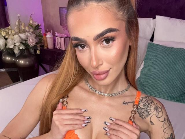 MarissaFex, con lunghi capelli biondi, sopracciglia folte e labbra piene, indossa un reggiseno di pizzo arancione, tiene le mani sui seni, ha tatuaggi sulla spalla destra e una collana d'argento..