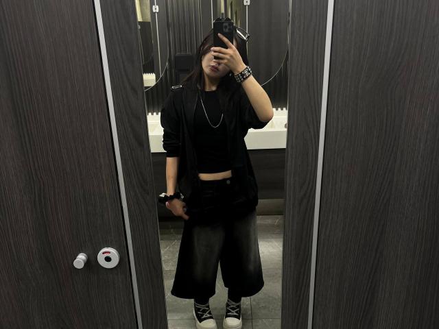 PeiBlank stands in a bathroom, wearing a black crop top, open jacket, black pants, and Converse sneakers, taking a mirror selfie with a camera, showing a wrist bracelet.