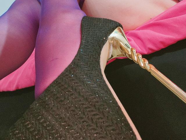 Akkanee's black, glittery high heel with a gold braid on the side is resting on her purple, sheer pantyhose-covered ass. Pink fabric is visible in the background.