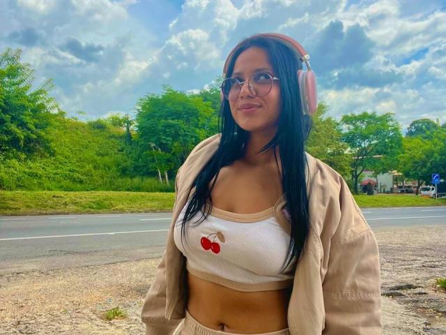 AlanaLuxe with long black hair, glasses, pink headphones, beige jacket, white crop top with cherry print, showing her tits, standing outside on a sunny day.