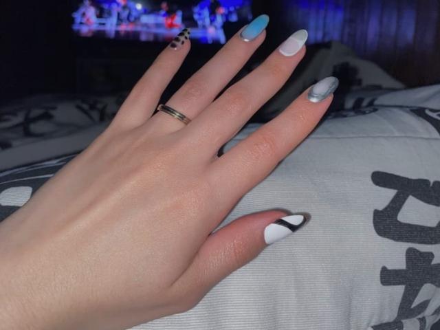SherylHill's hand with pale skin, wearing a gold ring, displaying long nails in blue, white, and black with a dot design. She's on a grey bed with white patterns.