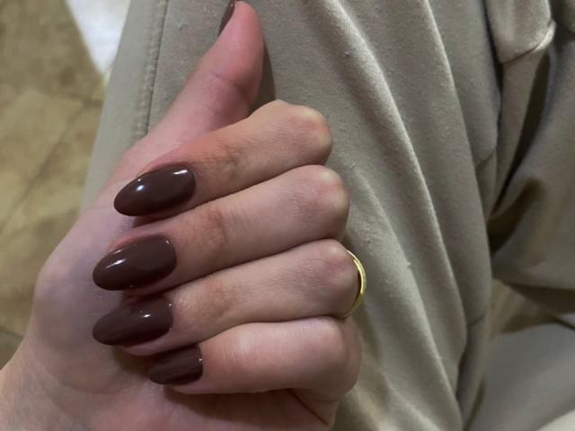SherylHill's hand with long, glossy brown nails, a gold ring on her middle finger, and beige pants. Her skin is pale, and the background is blurred.