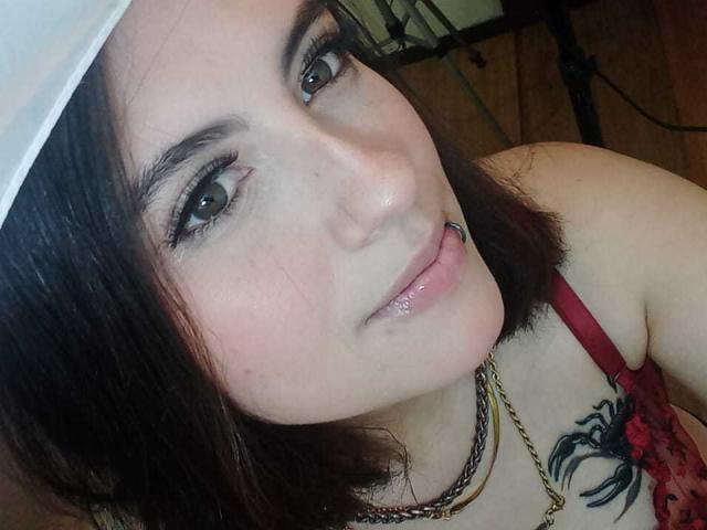 MaryCruzz with dark brown hair, green eyes, and a lip piercing, wearing a red lace top, chain necklace, and white cap, showing a black tattoo on her chest.