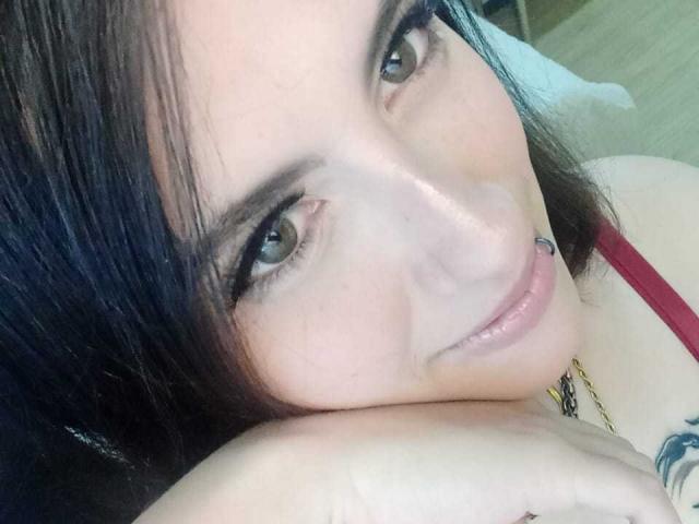 MaryCruzz's face, dark hair, green eyes, subtle smile, red tank top, nose piercing, light skin, resting chin on hand, small tattoo on shoulder, close-up selfie.