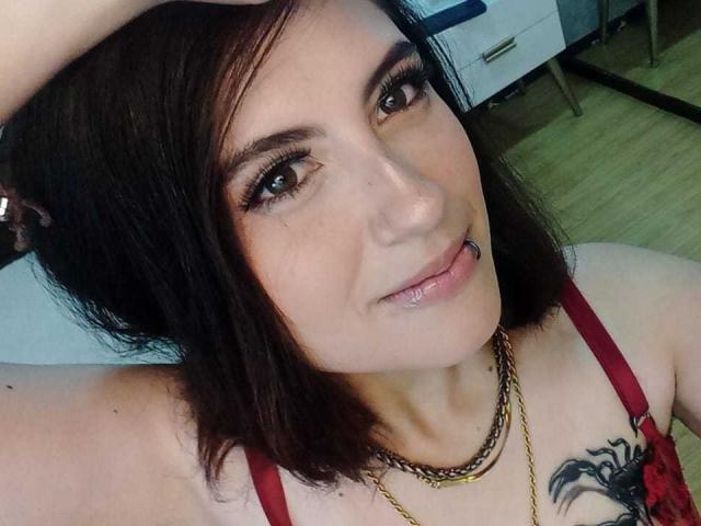 MaryCruzz has dark brown hair, brown eyes, a lip piercing, and a gold necklace. She's wearing red straps and has a black tattoo on her chest.