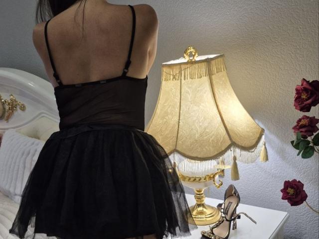 Rouzi wearing a black sheer top and black tulle skirt, back to camera, standing in front of a gold lamp with fringed shade, and red roses on a white table.