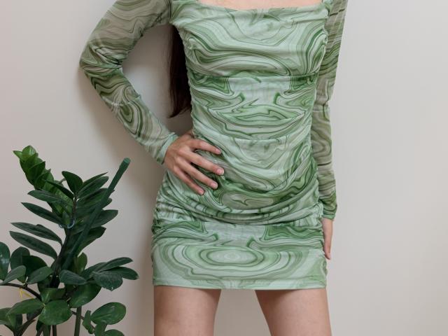 AmeliaMaiv in a green marbled dress, hand on hip, standing with a plant on the left, visible from neck to upper thighs, dress clinging to her tits and ass.