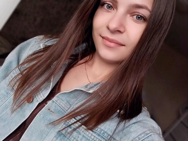 SaraUili with straight brown hair, blue eyes, light skin, and a subtle smile, wearing a light blue denim jacket, and a thin gold necklace.
