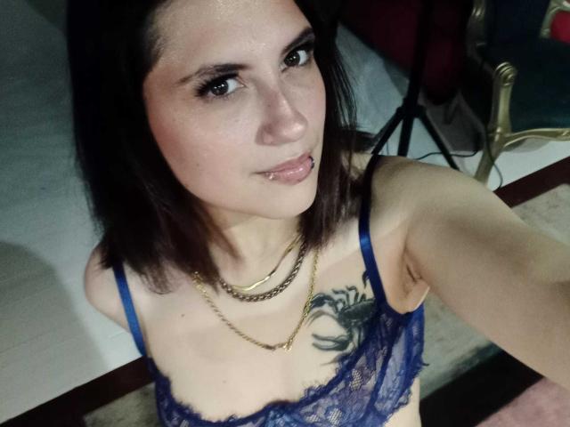 MaryCruzz, avec ses cheveux brun foncé, son soutien-gorge en dentelle bleu, son collier en or et son piercing aux lèvres, montre ses seins et son tatouage sur sa poitrine, et regarde l'appareil photo avec un léger sourire..