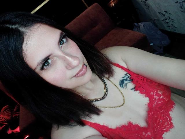MaryCruzz with pale skin, black shoulder-length hair, green eyes, red lace bra, small tits, gold chain, tattoo on right side, and slight pout, in dimly lit room.