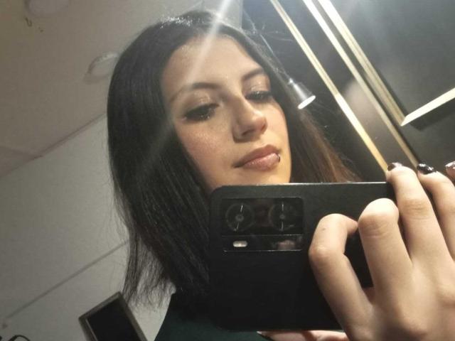 MaryCruzz, avec ses cheveux foncés, ses lèvres légèrement brillantes, tenant un téléphone noir, se prend en photo, montrant son visage et le haut de sa poitrine dans le miroir. Ses yeux sont à moitié fermés..