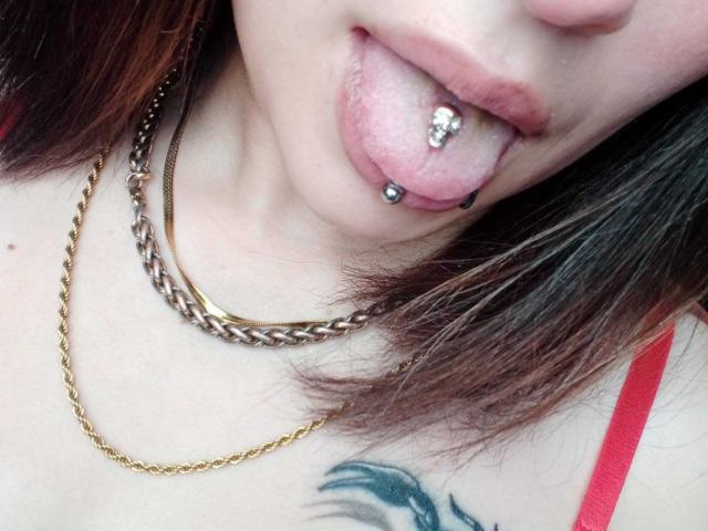 MaryCruzz's tongue is out with a gold chain and black ball piercing, showing a pink tongue with a silver jewel piercing. She's got a red strap and a black tattoo.