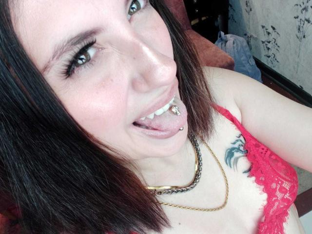 MaryCruzz with short brown hair, blue eyes, and a tongue piercing, sticking out her tongue, wearing a red lace bra, and a gold chain, showing a tattoo on her chest.