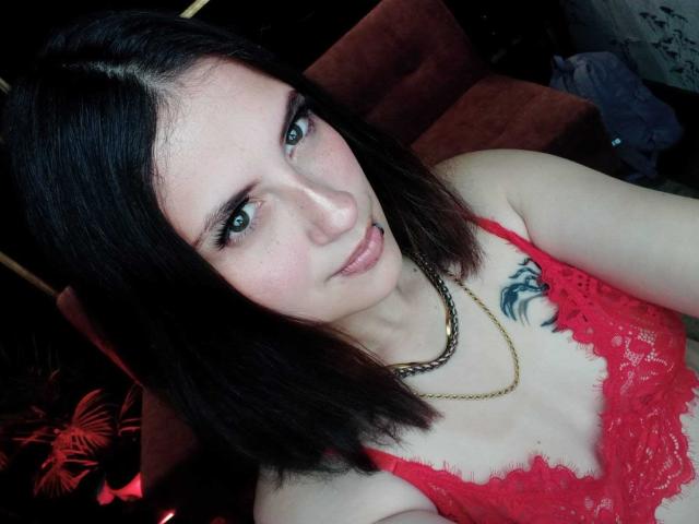 MaryCruzz with pale skin, dark straight hair, green eyes, red lace bra showing her small tits, gold chain necklace, black tattoo on right chest, subtle pink lips.