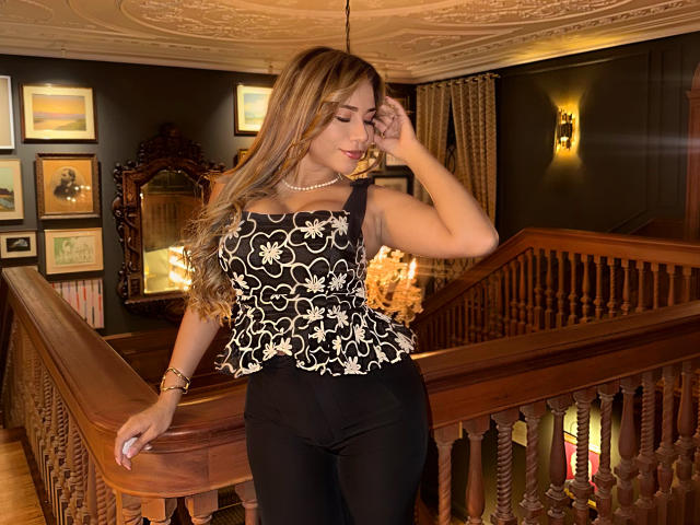 LunaMiranda with long blonde hair, black floral top, and black pants, standing on a wooden staircase, touching her face, wearing a pearl necklace, in a warmly lit room.