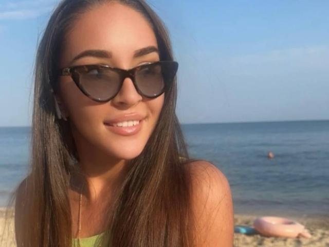 AquaLady with long brown hair, wearing black sunglasses, smiling at the camera, showing her white teeth, in a green top, on a sunny beach with a blue ocean.