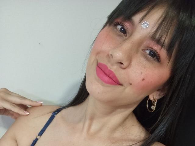 MadissonFontaine with pink lips, black hair, and a diamond forehead dot, wearing hoop earrings, and a blue strap, smiling with a small beauty mark on her cheek.