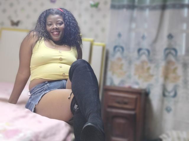 BrownieBabe with curly black hair, yellow crop top, denim shorts, and black thigh-high boots, smiling on a bed, showing her bare tan thighs and black boots.