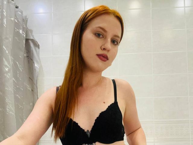 JannaCorsaro with red hair, black lace bra, pale skin, and pink lipstick, looking at the camera with a slight smirk. Her tits are small, and she's in a bathroom.