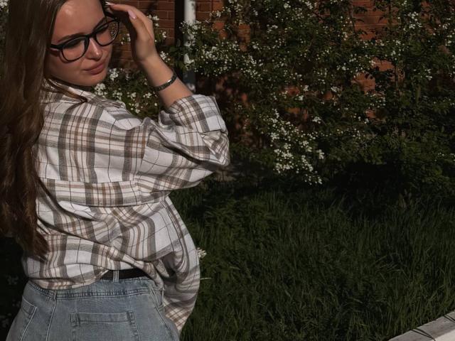 CatalinaBogar with glasses, long brown hair, plaid shirt, and blue jeans, adjusting her glasses, standing in sunlight, grass, and white flowers.