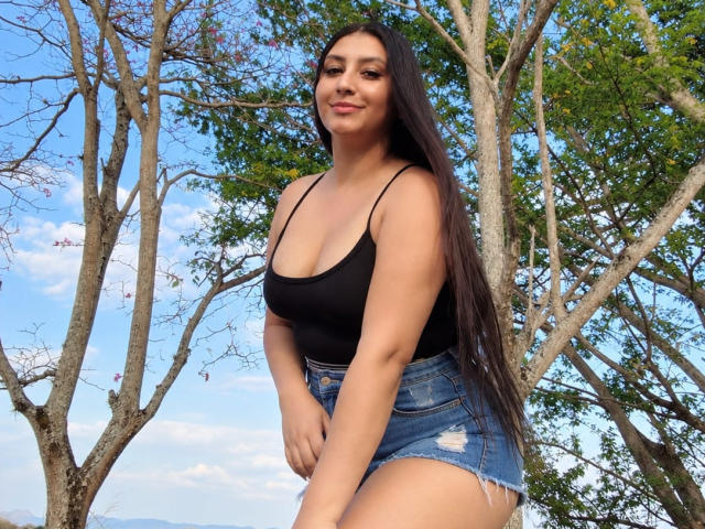 CatalinaRivero with long dark hair, black tank top, showing small boobs, and denim shorts, standing outdoors, smiling, arms slightly bent, blue sky and trees in background.