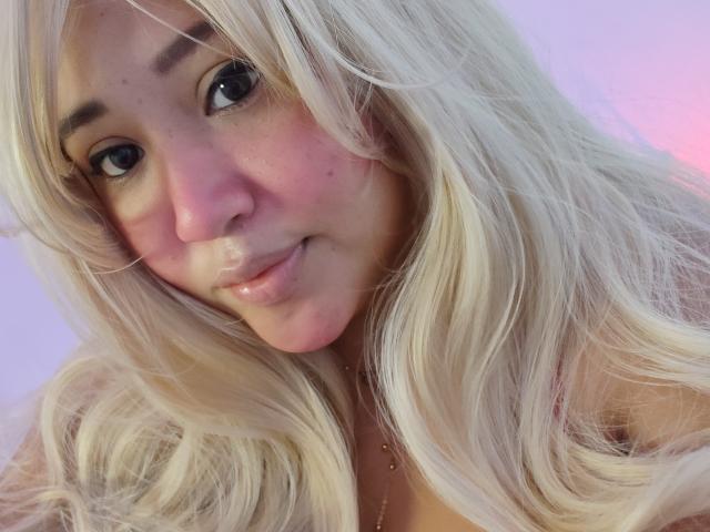 MafeFlor with blonde hair, pink cheeks, and glossy lips, showing a cute smile, has a small nose and dark eyes, looking at the camera with a slightly tilted head.