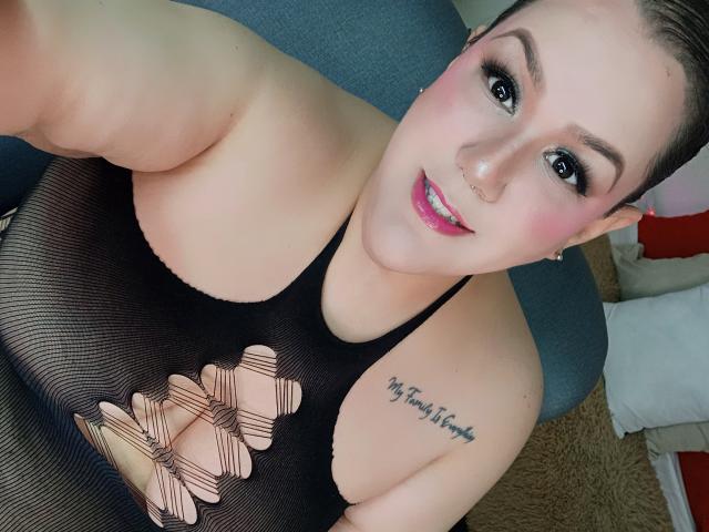GiiannaMorrys with pink lipstick, black mesh top showing her tits, tattoo on right shoulder, nose ring, and short brown hair, smiling at the camera.