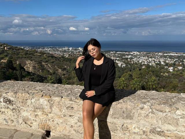 ShanitaMaud con vestido y chaqueta negros, sentada en un muro de piedra, sujetándose el cabello, sonriendo, cielo azul, nubes, océano, ciudad, colinas verdes al fondo, piel morena, gafas..