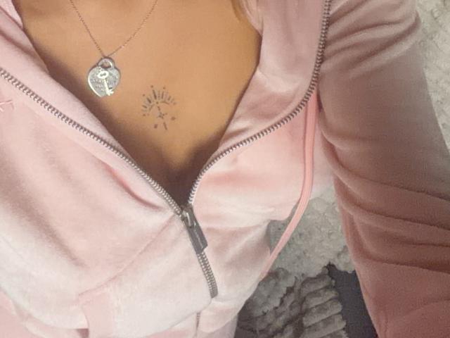 Il felponcino rosa con zip di SensualMila, che mette in mostra il suo décolleté con un piccolo tatuaggio di un sole tra le tette, indossa una collana d'argento con un ciondolo a forma di cuore..