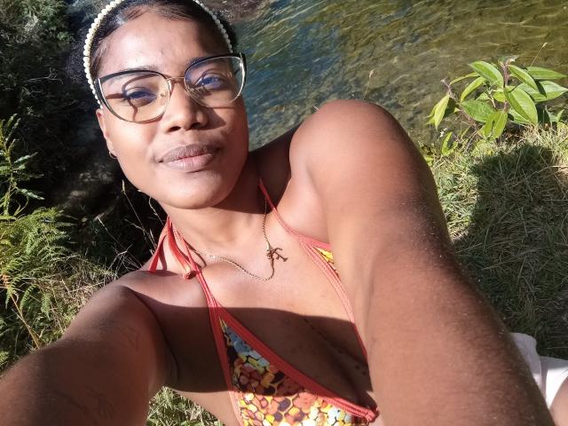 Caylin with dark skin, glasses, and a headband, wearing a red floral bikini, showing her small tits and a cross necklace, smiling by a clear, shallow water stream.