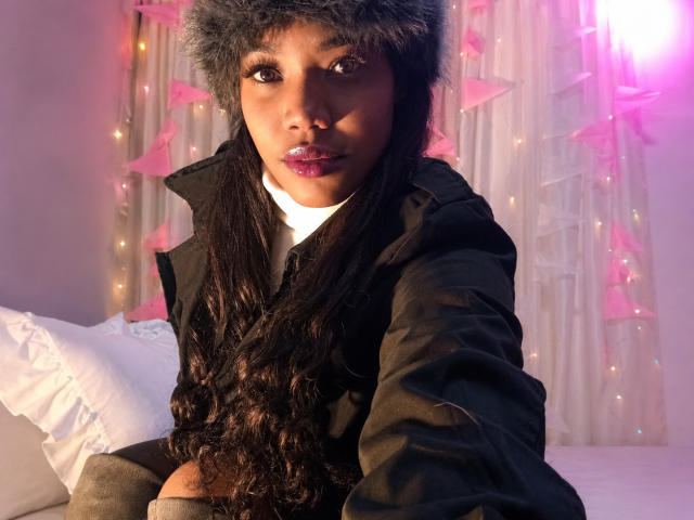 Caylin with long curly black hair, dark skin, and purple lipstick, wearing a fur hat and black jacket, sitting on a bed with white pillows, looking at the camera.