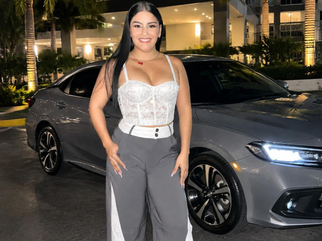 TalianaSquirt with long black hair, white lace bustier showing big tits, gray pants, standing in front of a shiny gray car, smiling at the camera.