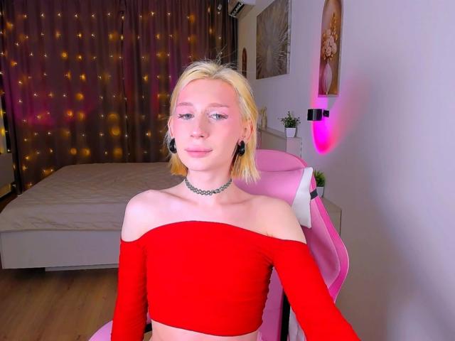 Mika-Candy-hot with blonde hair, red off-shoulder top, choker, black ear gauges, pink chair, bed, and wall-mounted art, showing a confident, cute, sexy, and attractive look.