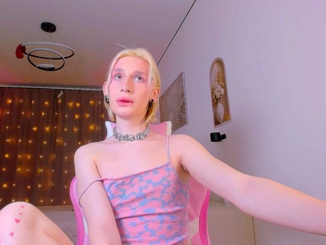 Mika-Candy-hot with pink and blue leopard print top, pink chair, silver choker, blonde hair, pink lipstick, sitting with legs spread, exposing knee with pink heart tattoo.