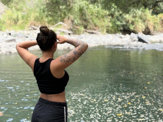 ThaliaAres with arms raised, black sports bra, tattooed arm, black shorts, standing by a river, water reflects light, green trees in background, short dark hair in a bun.