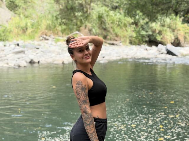 ThaliaAres with tattoos on her right arm, black sports bra, and black pants, standing by a river, hand covering her forehead, smiling, showing her ass.