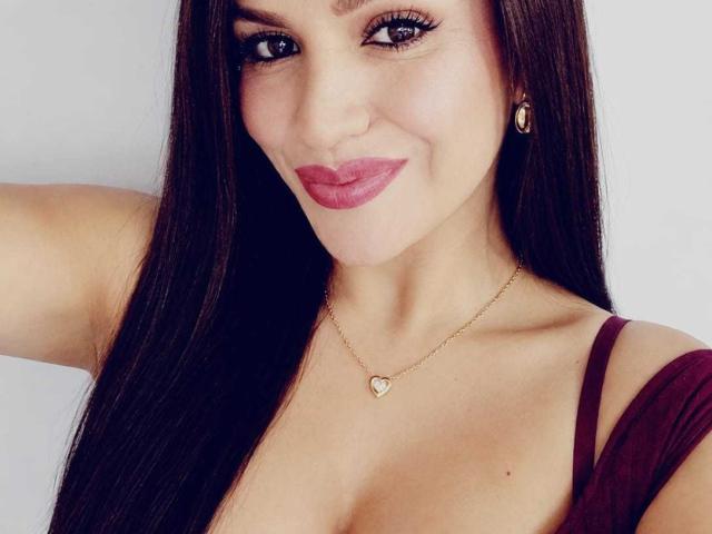 CameronViviane with long brown hair, wearing a maroon top, heart necklace, and red lipstick, smiling at the camera, showing her tits slightly. She's got cute earrings too.