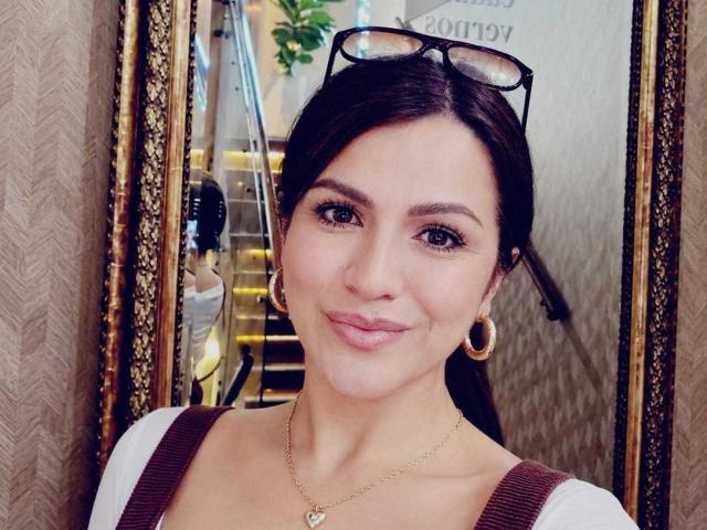 CameronViviane has dark brown hair, hoop earrings, and a gold necklace. She's smiling with pink lips, wearing a white shirt and brown vest, with glasses on her head.