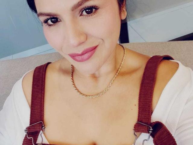 CameronViviane has light skin, brown eyes, and pink lipstick. She wears brown suspenders over a white top with a gold chain necklace. Boobs visible. Smiling.