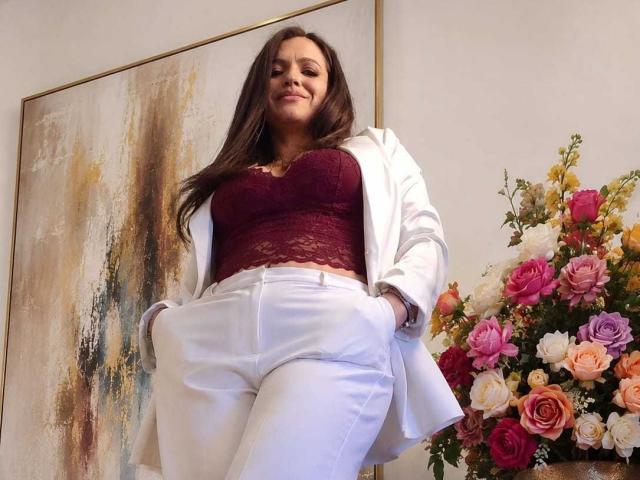 CameronViviane with long brown hair, wearing a maroon lace top, white pants, and a white blazer, hands in pockets, standing in front of a floral painting and bouquet.
