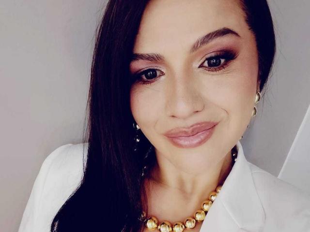CameronViviane with long black hair, dark eye makeup, pink lips, white shirt, gold necklace, earrings, and a slight smile. Boobs are not visible. She looks sexy.