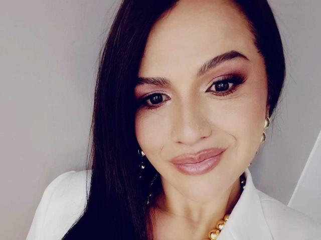 CameronViviane has dark, straight hair covering one eye, brown eyeliner, and pink lipstick. She's smiling with her tits barely visible in a white top, wearing gold earrings.