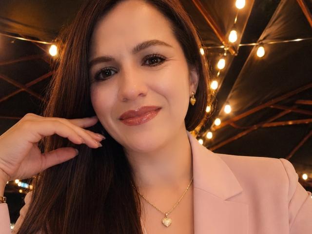 CameronViviane has shiny brown hair, glossy pink lips, and a cute smile. She's wearing a pink blazer, gold necklace, and earrings, with her hand near her face.